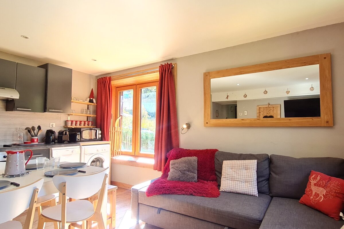 A bright, cozy studio apartment featuring an open-plan kitchen, dining area, and living space with a gray sofa, large mirror, and red curtains.