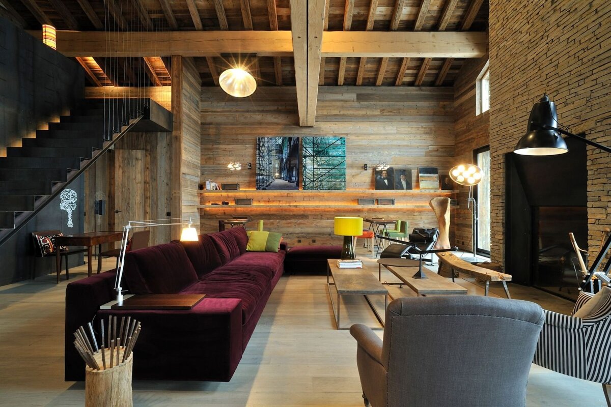 A rustic-modern living room features wood-paneled walls, exposed beams, a dark industrial staircase, and a long purple sofa, with a stone fireplace.