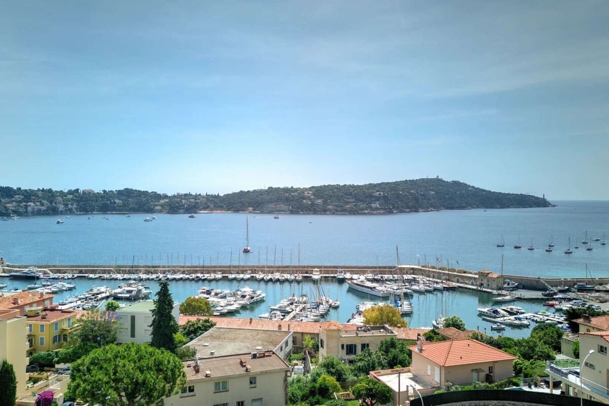 A scenic coastal town with a busy harbor full of boats and yachts. A blue bay extends to a distant green peninsula under a bright sky.