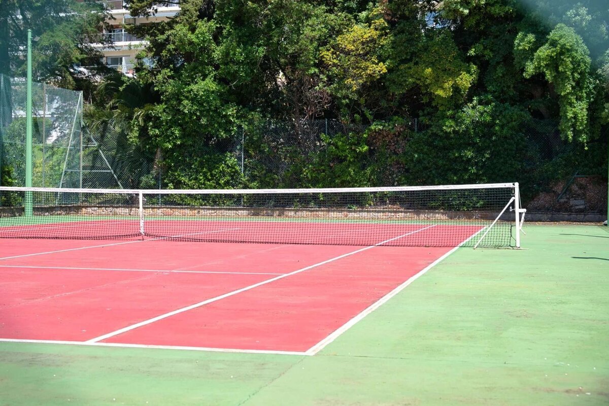 A vibrant red and green outdoor tennis court with a white net, backed by lush green trees and a glimpse of a building.