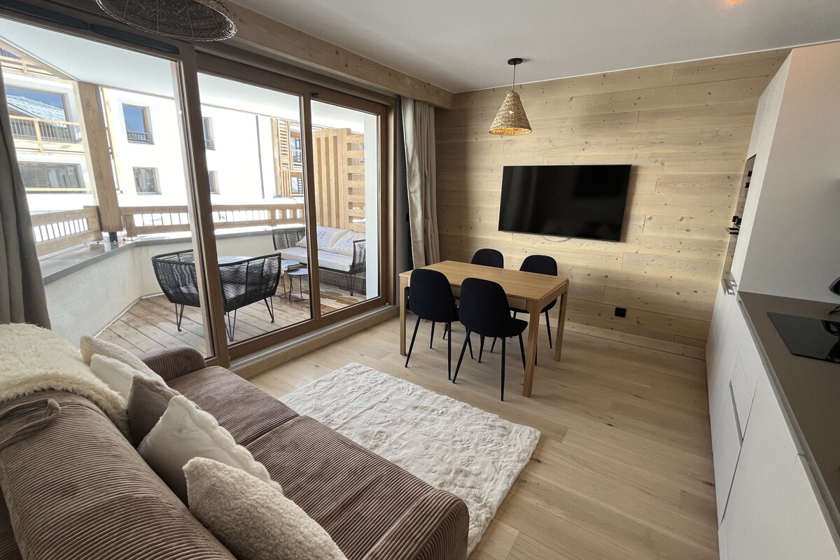 A cozy, modern living space features wooden walls, a dining area, TV, and a comfortable sofa. Large glass doors lead to a furnished balcony.