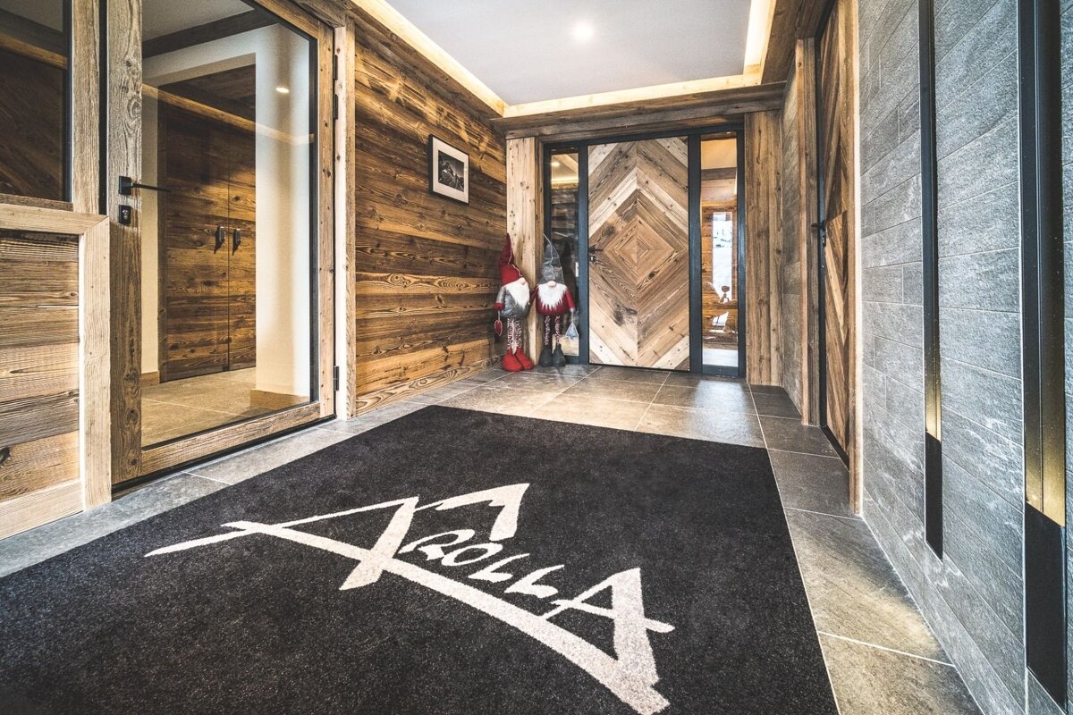 Rustic-chic chalet entryway featuring wood-paneled walls, a custom "Arolla" logo mat, and decorative gnomes near a chevron wooden door.