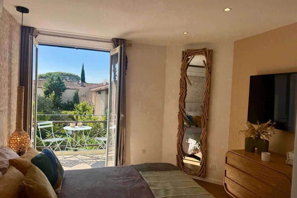 Cozy bedroom with a balcony overlooking green trees and houses. Features include a unique mirror, a dresser with a TV, and a comfy bed.