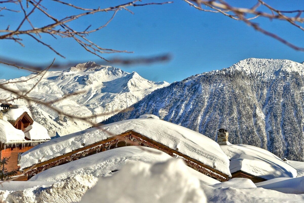 Winter wonderland: Snow-laden chalets nestled among majestic, white-capped mountains under a vibrant blue sky, with bare branches framing the view.