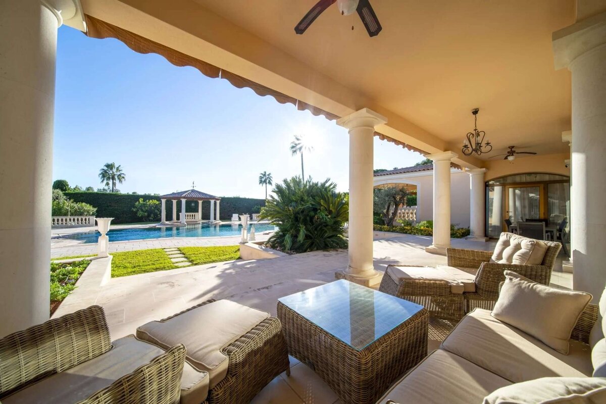 A luxurious covered patio with wicker furniture overlooks a sparkling pool, a gazebo, and palm trees under a bright sunny sky.