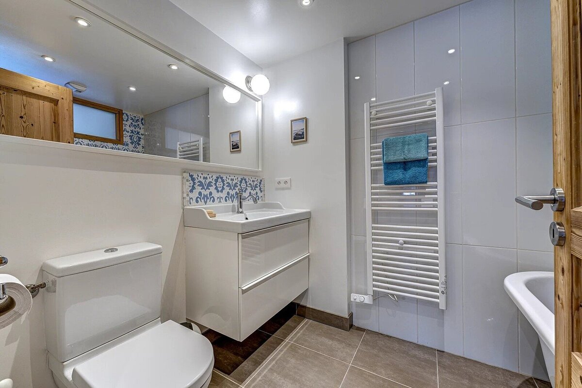 A bright modern bathroom featuring a white toilet, vanity sink with mirror, and a white heated towel rail with blue towels.