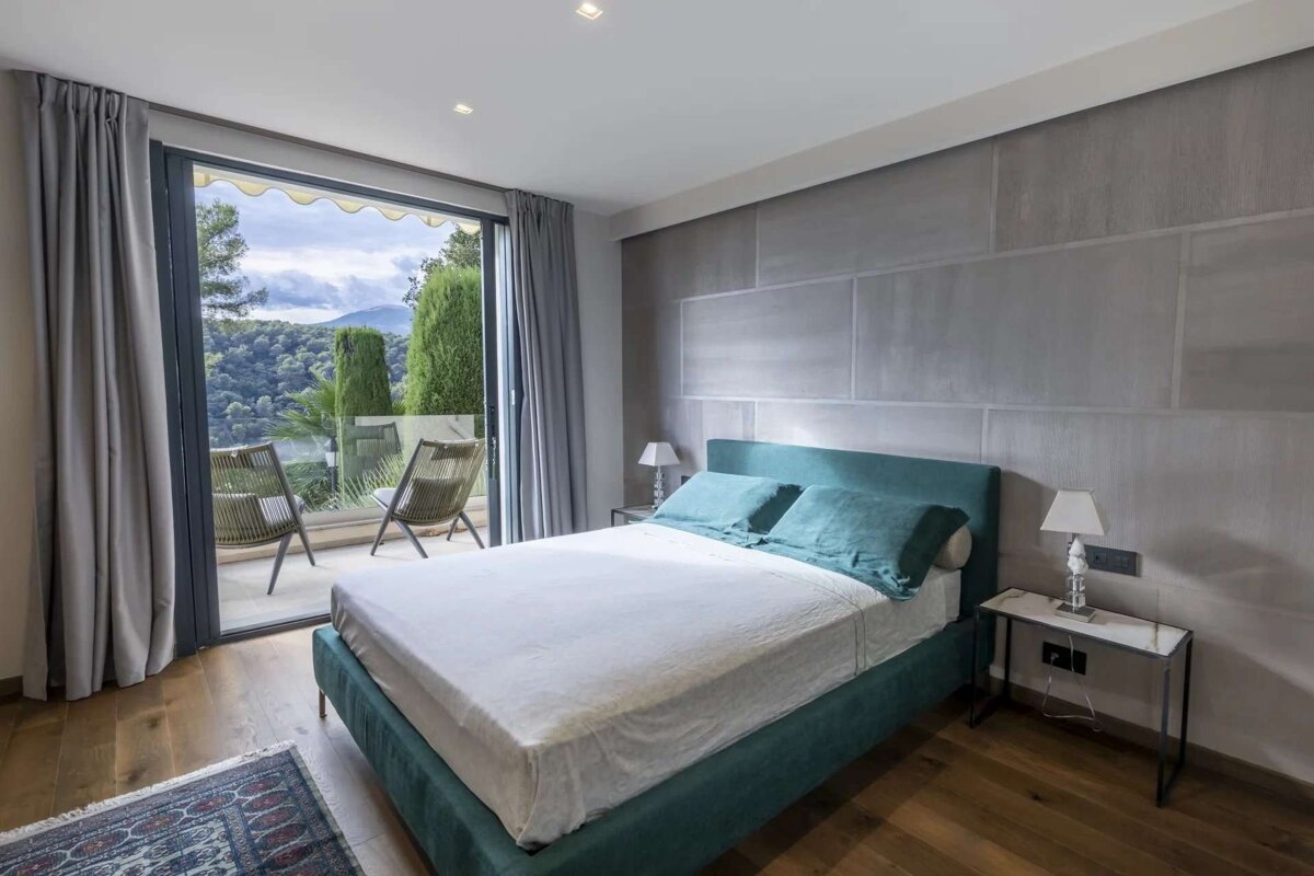 Modern bedroom with a teal bed, gray wall, and wooden floor. A balcony offers scenic mountain and forest views.