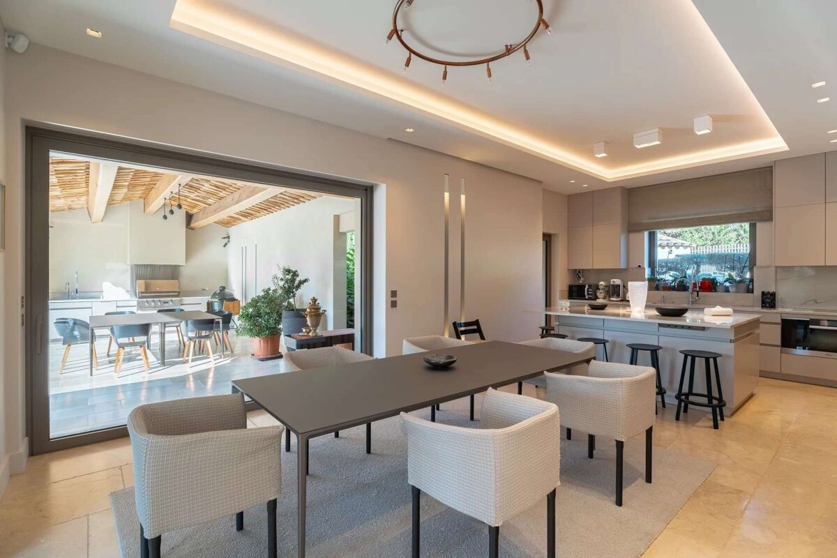 Modern open-plan kitchen and dining area with sleek design, recessed lighting, and large sliding doors opening to an outdoor patio with another kitchen.