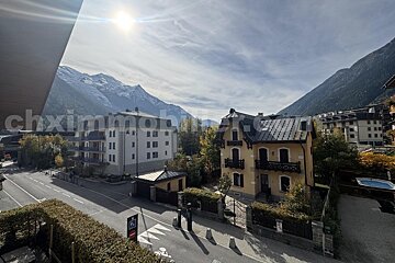 A sunny mountain town view featuring snow-capped peaks, a street, a modern apartment building, and a traditional yellow house with an ornate roof.