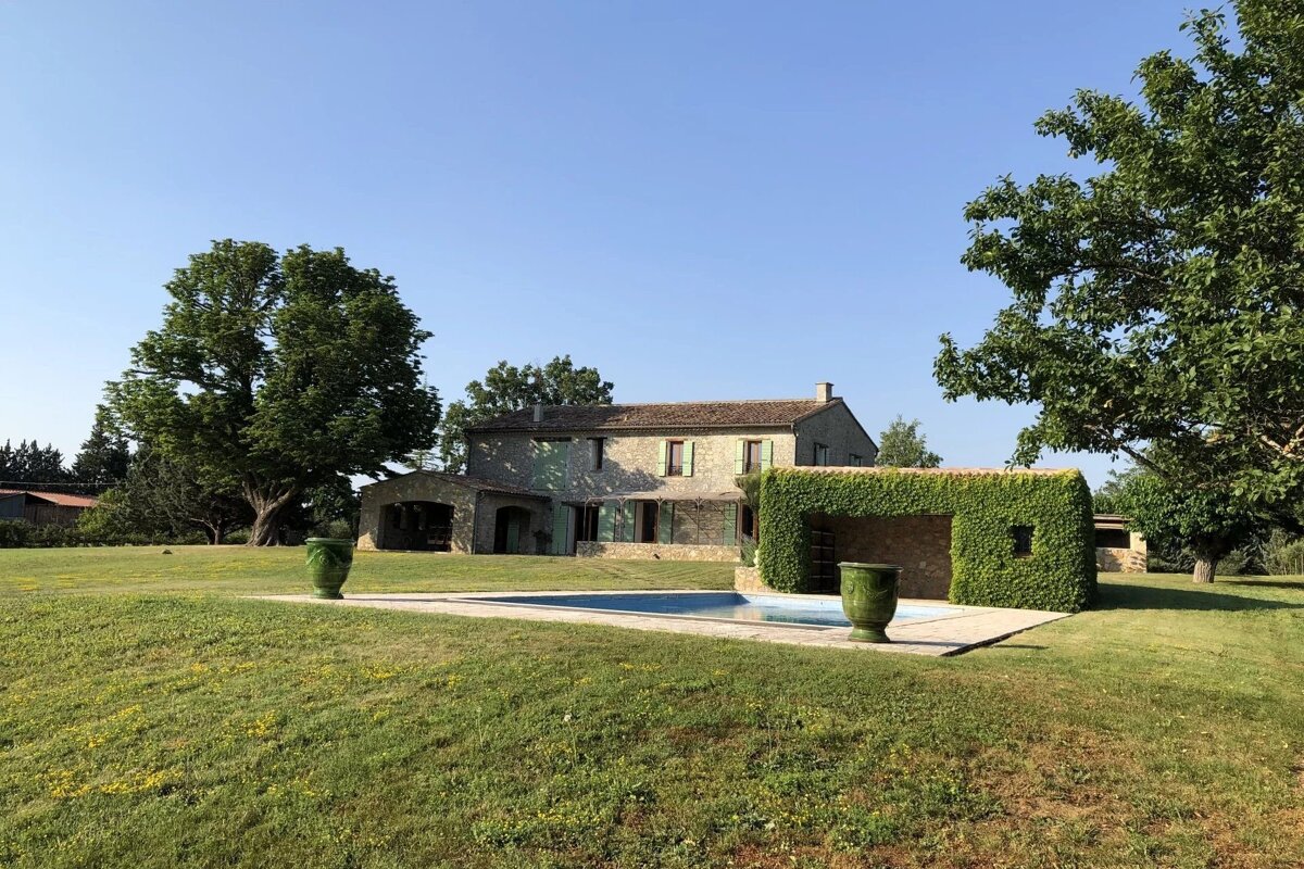 A stone house with green shutters, a swimming pool, and a lush green lawn, surrounded by trees under a clear blue sky.
