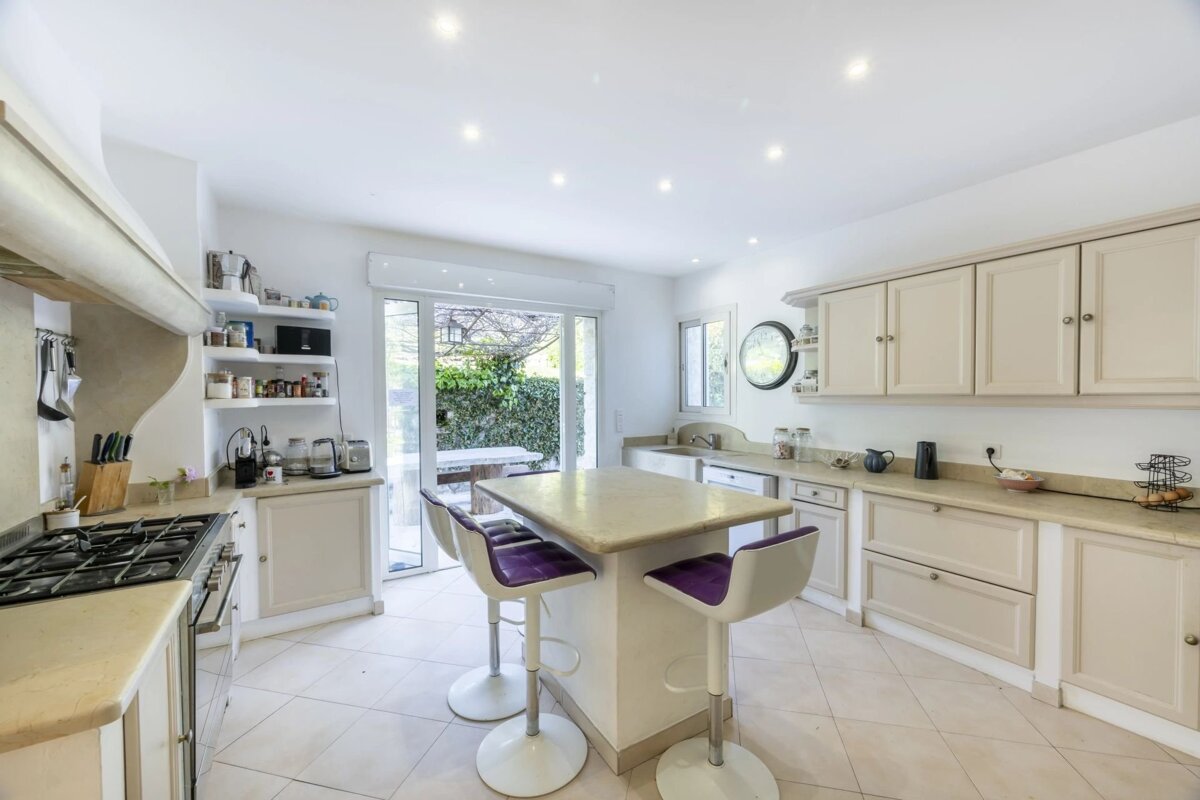 Bright kitchen with beige cabinets, a central island with purple bar stools, and a glass door leading to a garden.