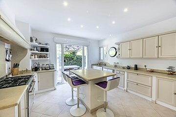 A modern beige kitchen featuring an island with three purple bar stools and a view of the outdoors through glass doors.