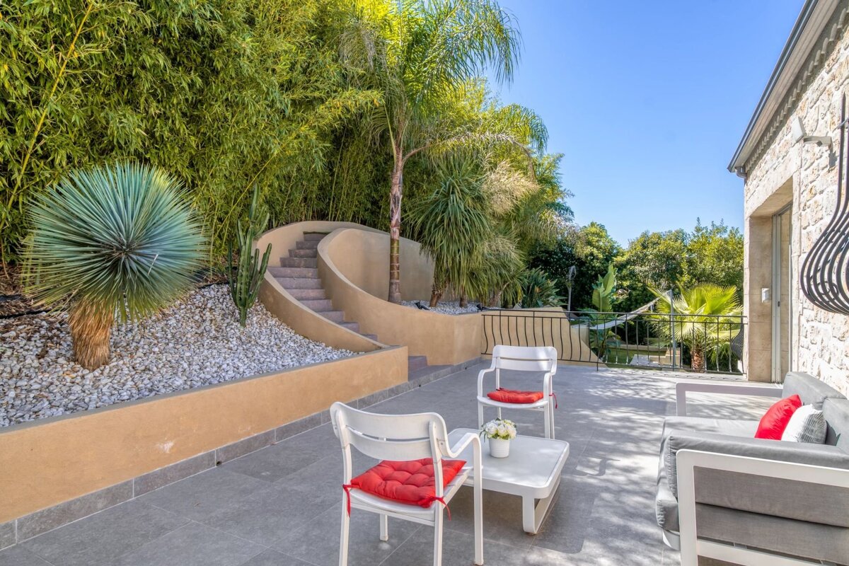 Sunny patio with modern white furniture, red cushions, and a curved staircase leading up to lush, tropical landscaping.