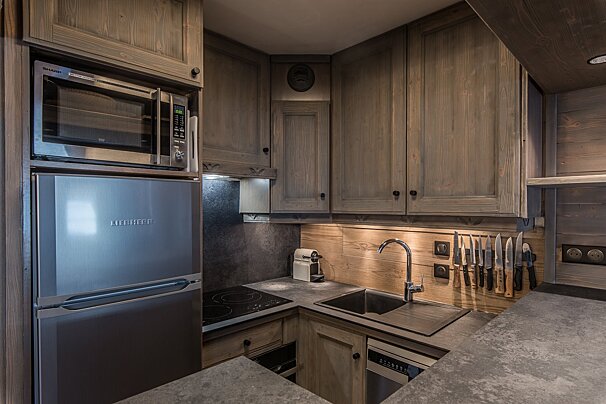 A compact, stylish kitchen with rustic wooden cabinets, stainless steel fridge, microwave, and stove, illuminated by warm under-cabinet lighting.