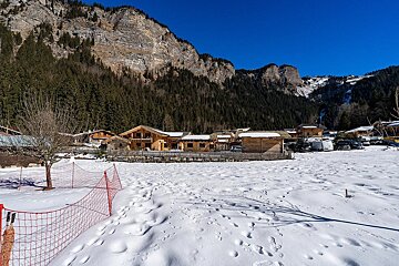 A snowy mountain village features rustic log cabins nestled against pine-forested peaks under a clear blue sky, with footprints marking the foreground.