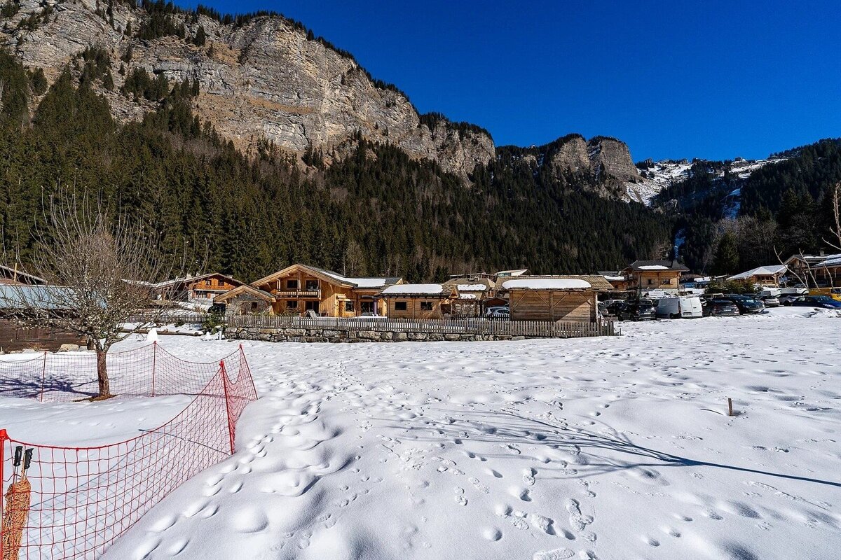 Wooden chalets nestled below massive rocky mountains in a snow-covered alpine village under a clear blue sky.