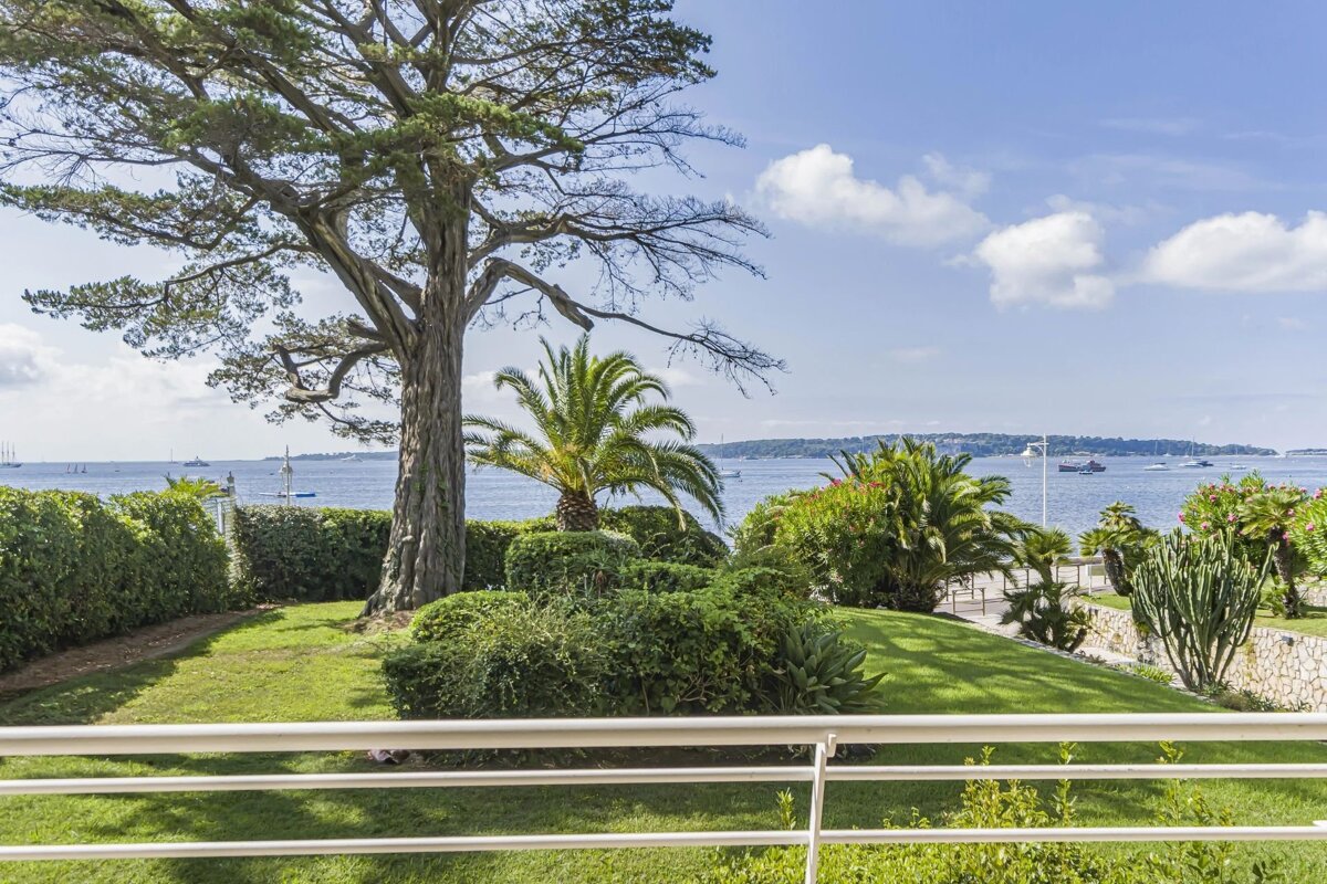 Lush garden with a large tree and palm trees overlooking a calm blue sea, viewed from a white balcony.
