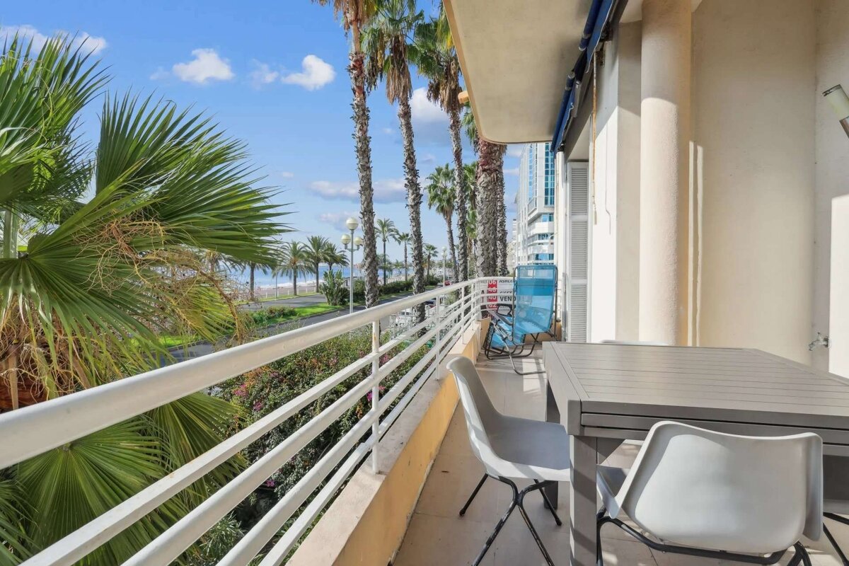 A sunny balcony overlooks a palm-lined promenade, beach, and ocean. It features an outdoor table, chairs, and distant buildings.