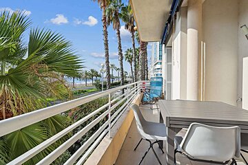 A sunny balcony overlooks a palm-lined promenade, beach, and ocean. It features an outdoor table, chairs, and distant buildings.