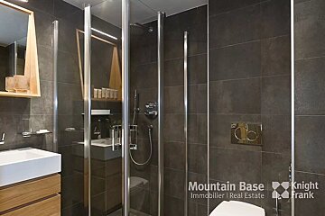 A modern bathroom features dark grey tiled walls, a glass shower enclosure, a white sink with a wooden vanity, and a mirror.