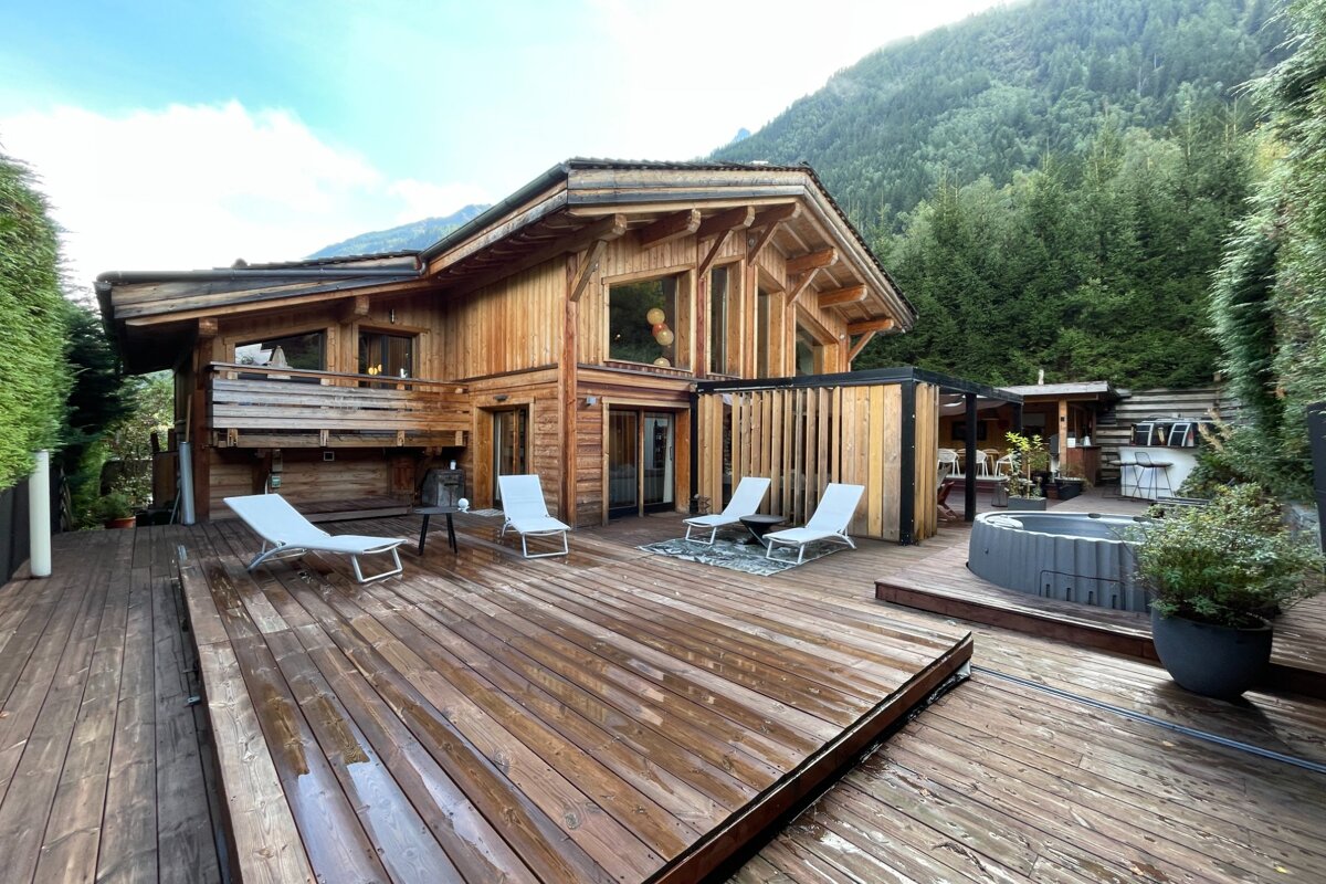 Rustic wooden chalet with a spacious wet deck, sun loungers, and hot tub, set against a verdant mountain and forest.