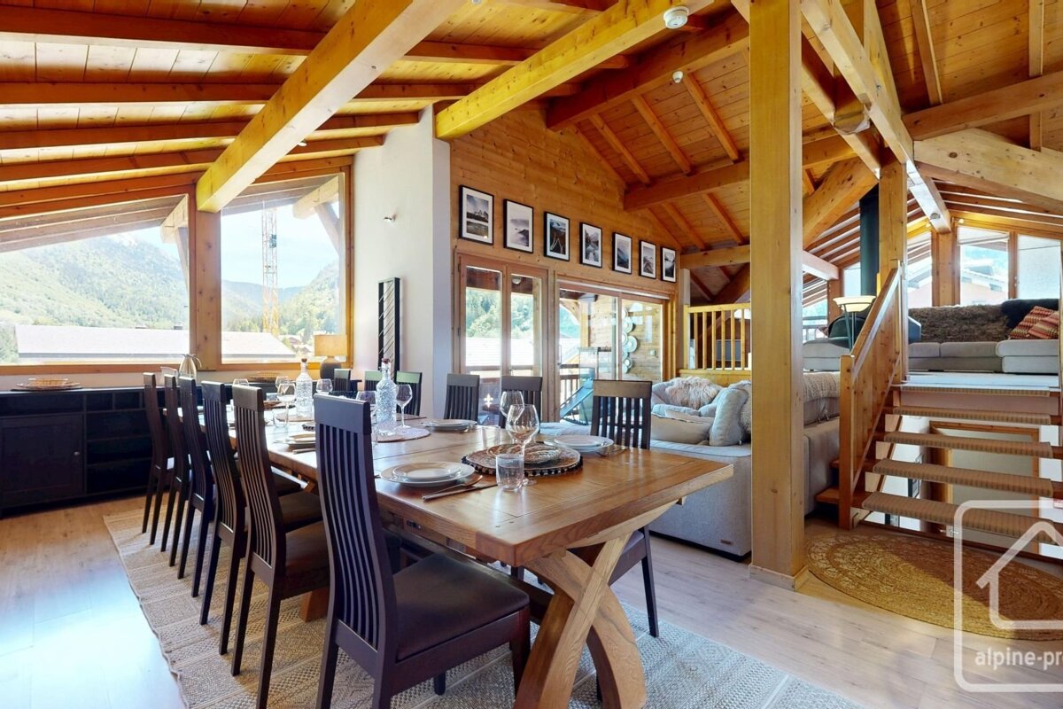 A spacious wooden cabin interior with a dining table, chairs, and stairs, offering mountain views.