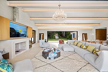 Bright, modern living room with a gray sectional, white rug, TV, fireplace, and chandelier, opening to a beautiful green garden.
