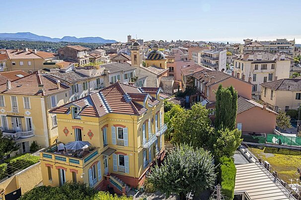 Aerial view of a charming yellow villa with blue shutters in a sunny, red-roofed Mediterranean town, mountains and sea visible.