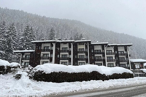 A large, snow-covered resort building nestled against a snow-laden pine forest on a mountain slope.