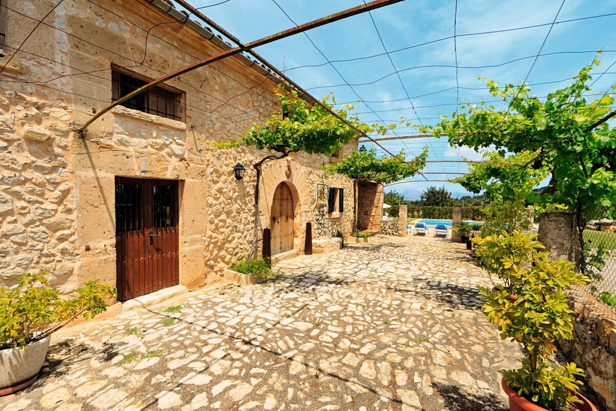 A charming stone building with a cobblestone courtyard under a leafy grapevine pergola. A swimming pool is visible in the sunny background.