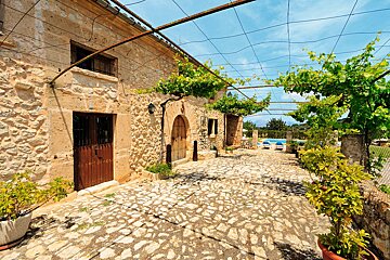 A charming stone building with a cobblestone courtyard under a leafy grapevine pergola. A swimming pool is visible in the sunny background.