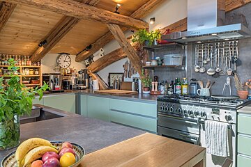 A warm, rustic kitchen featuring exposed wooden beams, light green cabinetry, and stainless steel appliances. Fruit and fresh herbs add life.