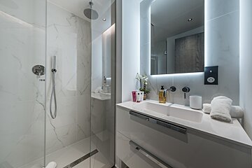 Sleek, modern bathroom with a glass shower, white marble walls, rain shower, illuminated mirror, and minimalist vanity with a white sink.