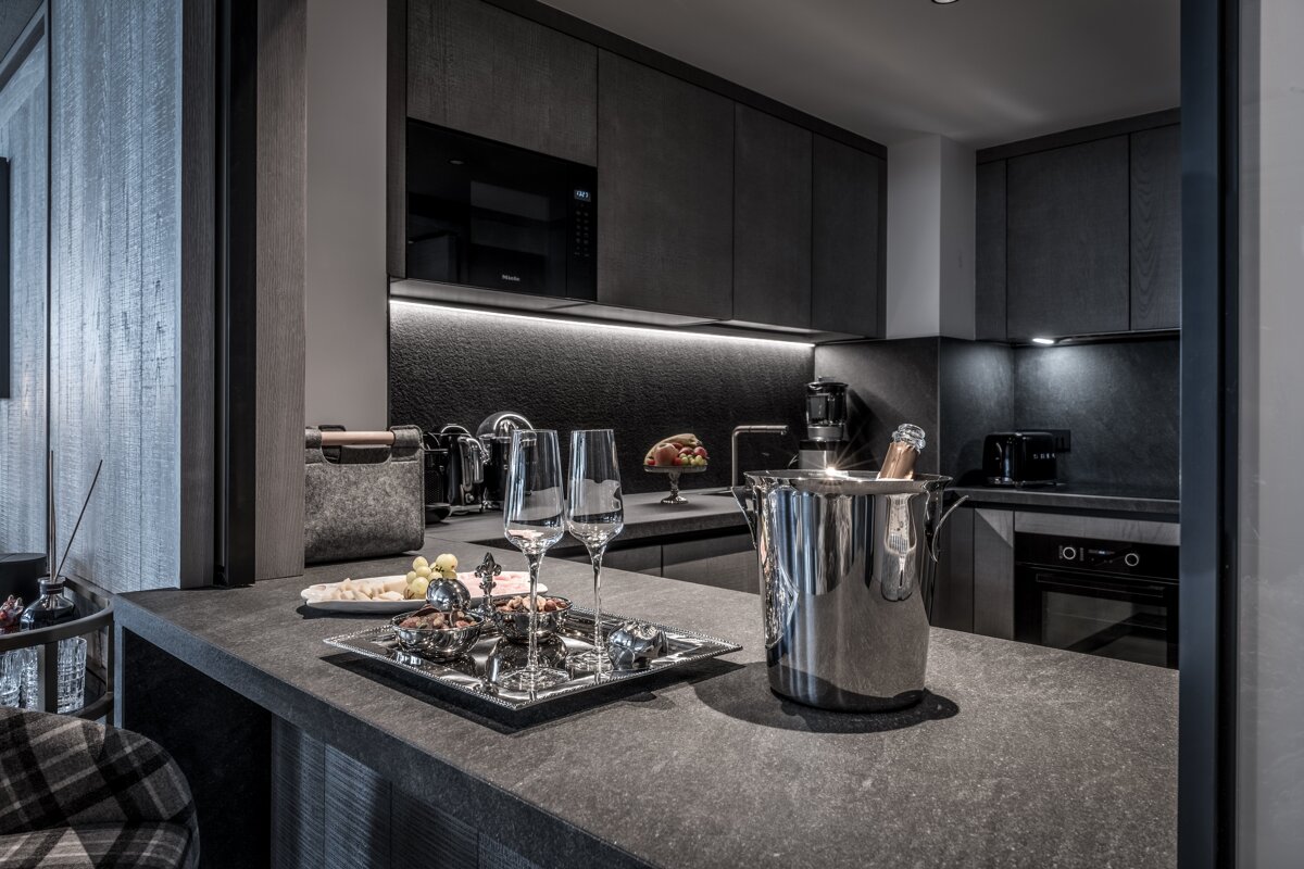A sleek, modern kitchen/bar area with dark cabinets, under-cabinet lighting, a champagne bucket with glasses, and snacks on a stone countertop.