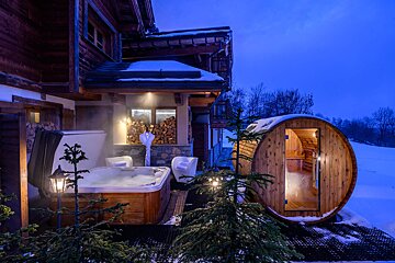 An outdoor hot tub and barrel sauna are set on a snowy deck at dusk, next to a rustic mountain chalet, creating a luxurious spa scene.