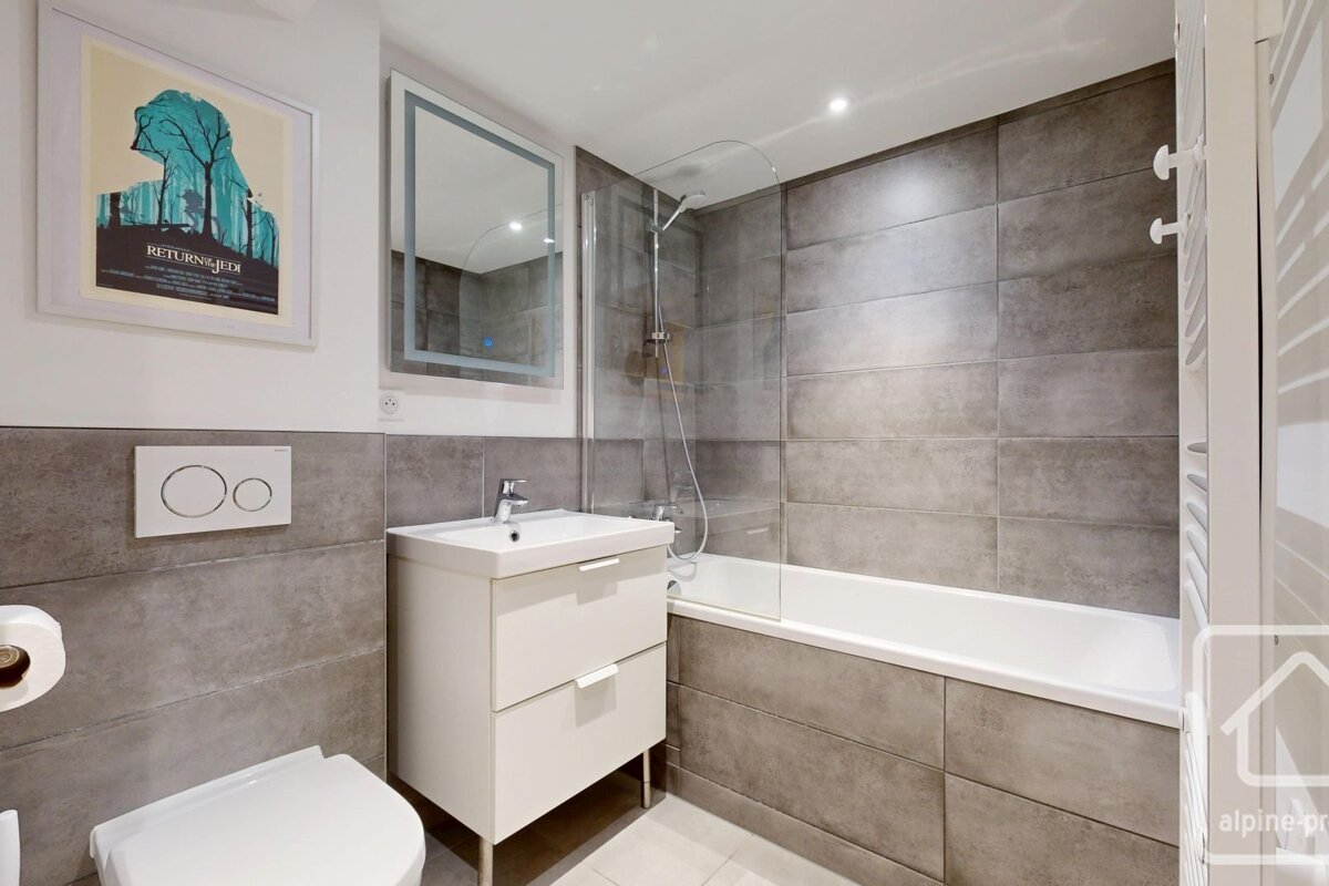 A modern bathroom featuring grey wall tiles, a white vanity and toilet, a bathtub with a glass screen, and a framed movie poster.