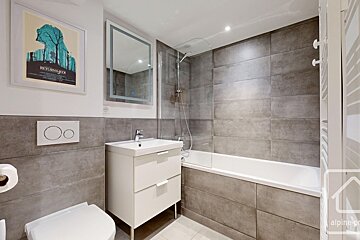 A modern bathroom featuring grey wall tiles, a white vanity and toilet, a bathtub with a glass screen, and a framed movie poster.