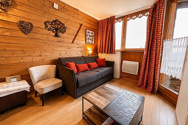 A cozy, wood-paneled living room featuring a dark grey sofa with red pillows, a beige armchair, and a rustic wooden coffee table.