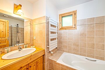 A warm-toned bathroom featuring beige tiled walls, a wooden vanity with a sink, a bathtub, a window, and a white towel warmer.