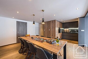Modern dining room featuring a long wooden table and grey chairs, opening to a sleek dark kitchen with wood accents.
