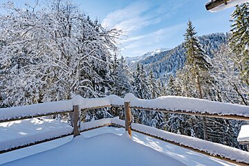 Sunlit, snow-covered mountain landscape with a wooden fence in the foreground, dense forest, and distant peaks under a bright blue sky.