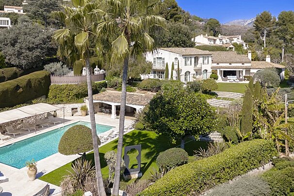 Aerial view of a sprawling white villa with a terracotta roof, large swimming pool, and lush terraced gardens, nestled on a sunny hillside with palm trees.