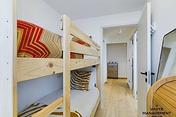 A bright room with wooden bunk beds featuring colorful bedding. A hallway with light wooden floors and a dresser is visible through an open doorway.