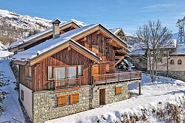 A rustic wooden chalet with a stone base and snow-covered roof nestled in a snowy mountain landscape.