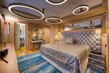 A modern bedroom with light wood walls, a large bed, gray tufted headboard, blue abstract rug, and striking circular ceiling lights.