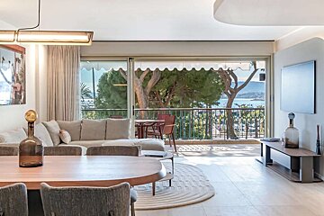 Modern living room with a sofa, dining area, and a balcony offering picturesque sea views framed by lush trees.