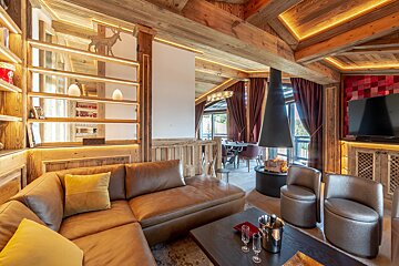 A luxurious chalet living room featuring rustic wood beams, a large brown sectional sofa, warm accent lighting, and a modern hanging fireplace, with a dining area.