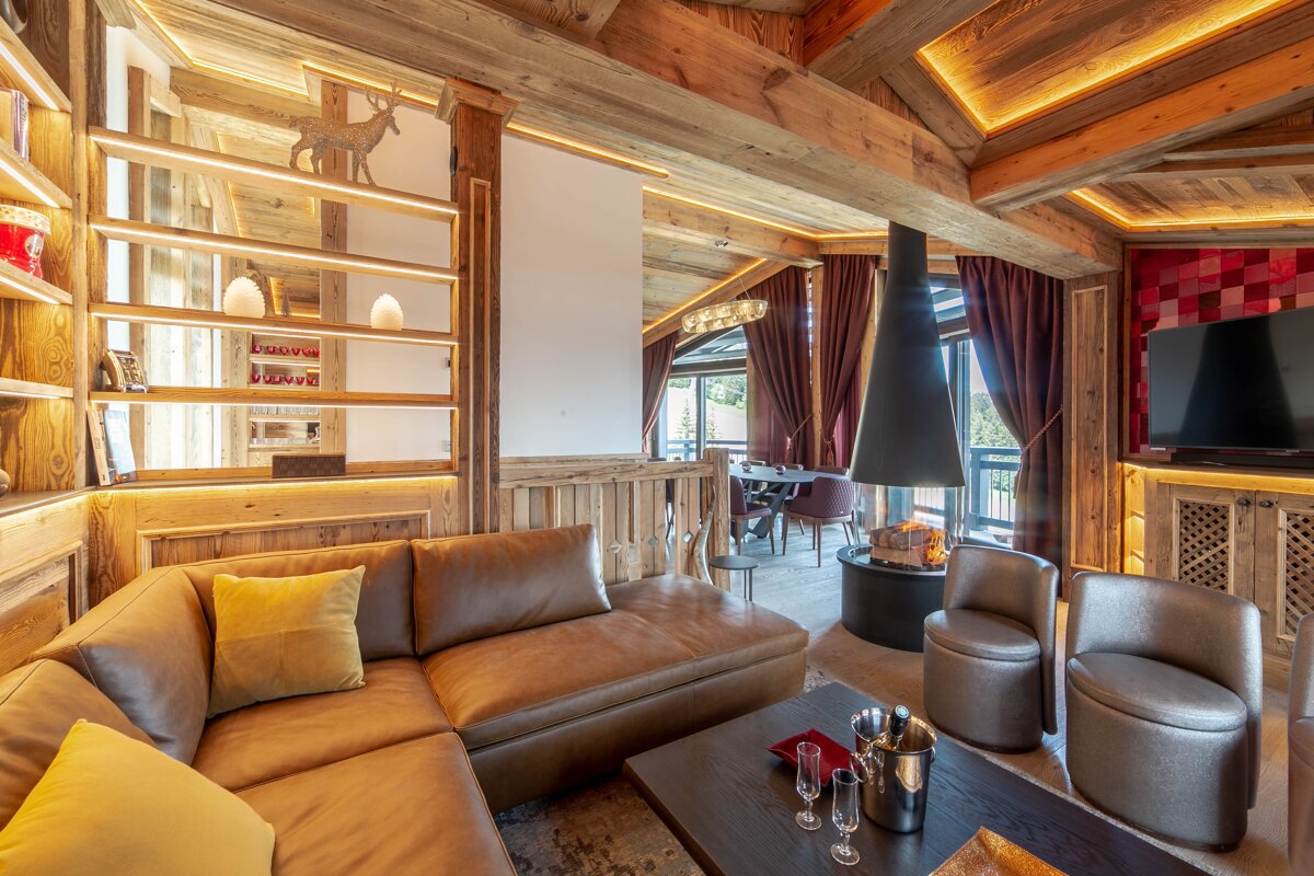 Cozy, modern chalet living room featuring rustic wood walls, a brown leather sectional, a sleek black fireplace, and warm lighting.