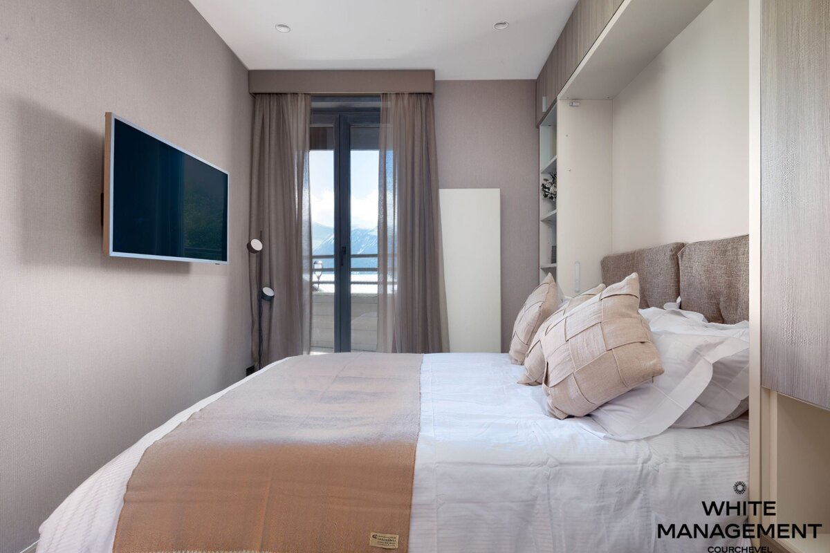 A serene, modern bedroom with a large bed, wall-mounted TV, and a balcony offering a snowy mountain view, all in neutral tones.
