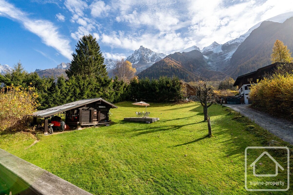Sunny alpine view of a bright green lawn, shed, and patio, framed by majestic snow-capped mountains and autumn trees under a blue sky.